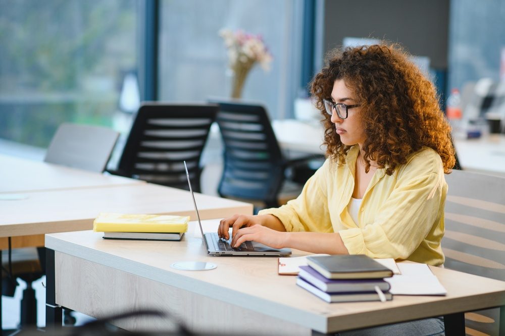 Studentin in gelbem Hemd arbeitet konzentriert an einem Laptop am Schreibtisch